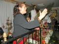 Una mujer con cabello largo y rizado, vestida de negro, se encuentra en una habitación decorada con velas y flores. La imagen muestra detalles de la decoración, como un árbol de Navidad y una figura religiosa en el fondo. La mujer parece estar en un momento emocional, posiblemente cantando o rezando.