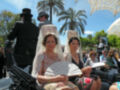 Dos mujeres vestidas con trajes victorianos y velos blancos, posando para una foto en un evento al aire libre con palmeras y cielo azul.