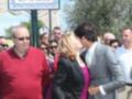 En el centro, una mujer con vestido rosa besa a un hombre en un evento público. A la izquierda, un hombre en rojo y a la derecha, otro en gris. Detrás de ellos, una placa con el nombre "Avenida Eva González" y un grupo de personas observando.