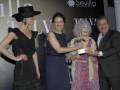 Una mujer en un vestido negro con sombrero de copa, una mujer con vestido azul y una anciana en un vestido floral reciben un premio de manos de un hombre con corbata. El fondo muestra una pantalla con el texto "Sevilla" y "Telva".
