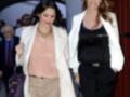 Dos mujeres caminan juntas, una con un blazer blanco y una camisa de color claro, la otra con un blazer blanco sobre una camiseta negra y pantalones negros. La imagen parece ser de una celebridad en un evento formal.