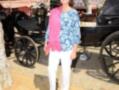 A una mujer vestida con un top de flores azules y blanco, pantalones blancos y una falda rosa. Está en un entorno con carros de caballo, posiblemente en una feria o evento cultural.