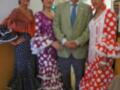 Una imagen de tres mujeres vestidas con trajes típicos flamencos, posando junto a un hombre. La escena parece ser una celebración cultural o festiva en España, con decoraciones de azulejos y flores al fondo.