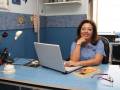 Profesora sonriendo en su escritorio, con laptop y libros al lado, en un ambiente de oficina.