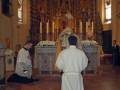 Imagen de un altar en una iglesia, con dos sacerdotes rezando frente al altar. El altar está decorado con flores y una imagen central. La iglesia tiene vitrales en las ventanas laterales.