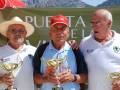 Tres hombres celebran con trofeos en un evento al aire libre, con un fondo de montañas y un letrero que dice 'Puerta de la Sierra'.