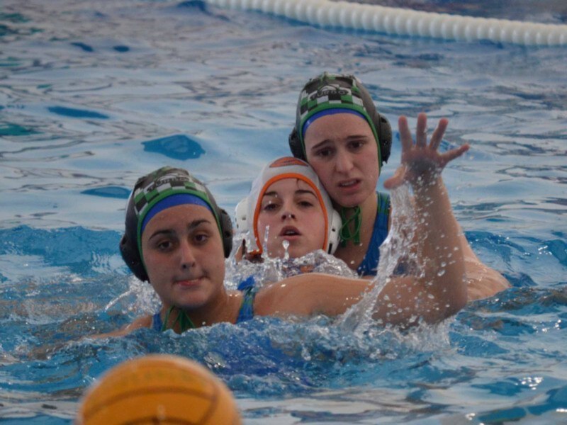 Tres jugadoras de waterpolo en acción, con cascos y una pelota amarilla flotando en el agua.