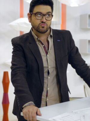 Un hombre con una camisa beige y un chaleco negro, posando frente a una mesa blanca en un entorno moderno con estanterías y lámparas.