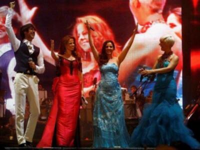 Tres personas cantando en un escenario con vestidos de gala, uno en blanco y dos en azul, con un fondo de pantalla con imágenes de personas sonrientes.