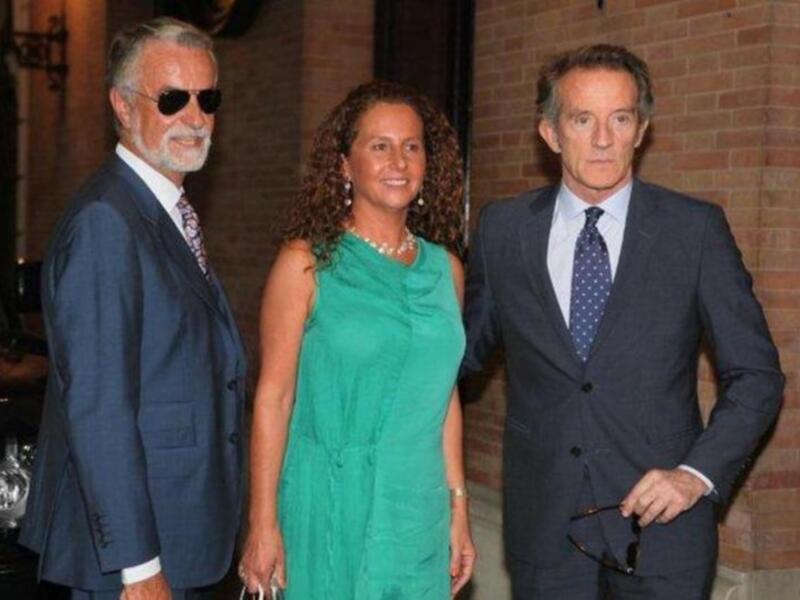 Tres personas elegantes posando juntas al aire libre, vestidas formalmente, con un fondo de ladrillo y un coche de época en el lado izquierdo.