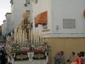 Imagen de una procesión religiosa con una imagen de la Virgen en el centro, rodeada por velas y flores. La imagen se encuentra en un edificio blanco con una placa de mención. Personas observan la procesión desde el lado derecho.