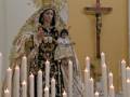 Imagen de una Virgen con un niño en sus brazos, vestida con trajes tradicionales y rodeada de velas encendidas. En el fondo, un Cristo crucificado en una cruz y una pared amarilla.