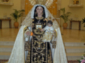 Imagen de una Virgen María vestida con un manto negro y dorado, rodeada por flores y adornos. En el fondo, se pueden ver otros santos en estatuas.