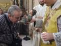 Un sacerdote sostiene una imagen religiosa mientras interacciona con una persona que sostiene una cámara en una iglesia.