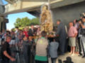 Una imagen de una procesión religiosa con una estatua de la Virgen en el centro, rodeada por fieles y fotógrafos. El cielo es azul claro y hay árboles al fondo.