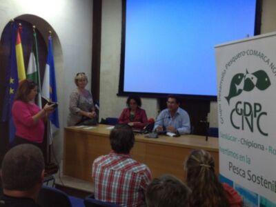 Una persona toma una foto mientras otros dos hablan en un escenario con banderas y un panel informativo.
