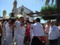 Un grupo de personas en uniforme naval participa en una procesión religiosa. La imagen muestra un altar con flores y una estatua, mientras el público observa la ceremonia. La escena se desarrolla en un entorno urbano con árboles y una estructura de madera.