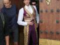 Un hombre vestido con un traje de baile tradicional posa junto a otros dos hombres, frente a una puerta de madera con detalles dorados.