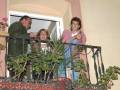 Un hombre y una mujer son vistos en un balcón, con plantas en el frente. La imagen sugiere una celebración o reunión familiar.