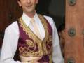 Adrien Brody con un vestido tradicional de color morado y dorado, posando frente a una puerta de madera.