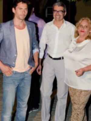 Tres personas posando juntas en una fiesta nocturna, dos hombres y una mujer. El hombre de la izquierda lleva un blazer azul claro, el hombre del medio está con una camisa blanca y pantalones grises, mientras que la mujer de la derecha lleva una camisa blanca y pantalones con estampado animal.