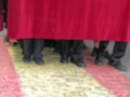 Pié de foto: Detalle de piernas con traje negro y falda roja, posiblemente en una ceremonia o evento formal.