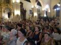 Una multitud de personas en una iglesia, con arcos y vitrales. La imagen muestra a varias mujeres sentadas en filas, algunas con joyería y vestidos elegantes. La decoración interior incluye estatuas religiosas, vitrales y una gran imagen central.