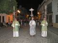 Una procesión religiosa nocturna con tres personas vestidas con hábitos blancos y verde, cada uno sostiene un bastón ceremonial. La escena se desarrolla en una calle adoquinada, con edificios blancos y luces de calle que iluminan la noche.