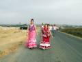 Dos mujeres caminan por una carretera, una con un vestido de flamenca rosado y la otra con uno rojo. El paisaje es desértico, con un camino asfaltado y una zona de basura en el fondo.