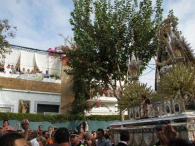 Una procesión religiosa con una imagen de la Virgen en un carro adornado con flores y decoraciones. La gente observa desde los balcones, mientras el carro pasa por una calle con árboles y edificios en el fondo.