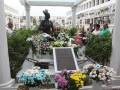 Una estatua de bronce en un cementerio, rodeada por flores y gente observando.