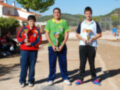 Tres jóvenes posan con trofeos en una escena al aire libre, con un paisaje de montañas y edificios en el fondo.