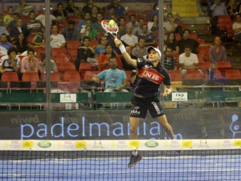 Jugador de pádel en acción durante un partido en una cancha cubierta, con público en el fondo.