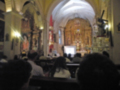 Interior de una iglesia con altares, retablos y un gran vitral en el fondo. Personas sentadas en las bancas observando algo en la pantalla frontal del altar.
