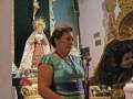 Una mujer en una iglesia, frente a un altar con una imagen de la Virgen María y un crucifijo. La mujer lleva una camisa verde con rayas y un vestido azul con cinturón. La imagen está en una iglesia con detalles dorados y una decoración festiva.