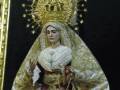 Imagen de una Virgen María vestida con trajes elaborados, posiblemente en un altar o escenario religioso. La imagen muestra la Virgen con una vestimenta ricamente bordada y un velo que cubre su rostro. La postura de la Virgen, con una mano levantada en gesto de bendición, sugiere un acto de veneración o adoración. El fondo es oscuro, lo que destaca la figura y su vestimenta dorada y plateada.