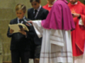 Un niño en uniforme negro sostiene un libro mientras un sacerdote en vestido morado y otro en chasuble roja lo observan.