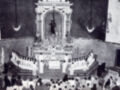 Imagen en blanco y negro de una iglesia con un altar central. En el fondo, se ve a un sacerdote en una posición de oración. La imagen muestra a personas sentadas en dos filas, con un grupo más numeroso en la parte frontal. La decoración del altar es elaborada y contiene elementos religiosos.