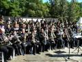 Una banda militar en uniformes oscuros se toca al aire libre con instrumentos como trompetas, tubas y flautas. El grupo está sentado en sillas al aire libre con un fondo de árboles y una estructura metálica.