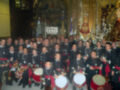 Una agrupación de personas vestidas con uniformes militares posando en una iglesia. La imagen muestra a varias personas, algunos con instrumentos musicales como tambores y trompetas. La decoración interior de la iglesia es elaborada, con altares y vitrales.