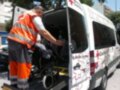 Un hombre en un chaleco naranja y pantalones de trabajo está ayudando a empujar una silla de ruedas hacia un ambulancia de Cruz Roja. La imagen muestra la acción de una persona ayudando a alguien en situación de necesidad, enfocándose en el trabajo colaborativo y la ayuda mutua.