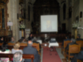 Interior de una iglesia con personas sentadas en bancos, un hombre hablando frente a una pantalla proyectada y flores decorando la pared.