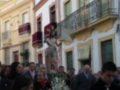 Una procesión religiosa en una calle de la ciudad, con personas vestidas formalmente y un crucifijo en el centro.