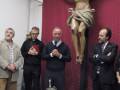En una iglesia, cuatro hombres observan con reverencia a un Cristo crucificado en la pared. En el centro, una mesa blanca sostiene un candelabro y flores blancas. La atmósfera es serena, con la iluminación suave y el rojo de la pared contrastando con los tonos más calidos.