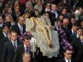 Una procesión religiosa con fieles vestidos de negro, seguidores de la Virgen María. La imagen central es una representación de la Pietà, la figura de la Virgen María cargando el cuerpo de Cristo muerto. La procesión ocurre al aire libre, con un cielo nublado y una atmósfera serena.