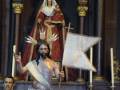 Una estatua de una figura religiosa, probablemente un santo, vestida con un manto rojo y tocada con una corona. La estatua sostiene una bandera blanca y un cetro dorado, mientras que una persona con corbata roja la observa desde detrás. La escena se desarrolla en un entorno arquitectónico con columnas doradas y una estructura de madera.
