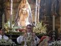 Imagen de una Virgen María en un altar, rodeada por flores y un sacerdote rezando.