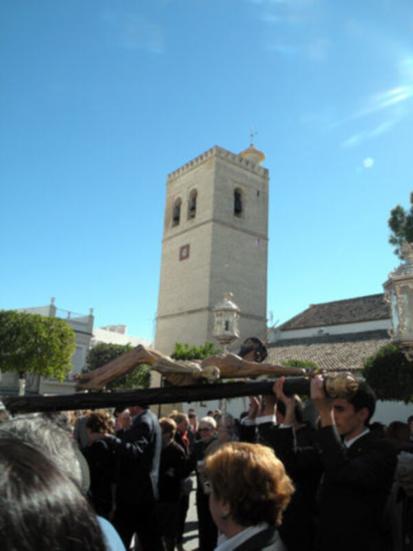 Una procesión religiosa con una cruz grande, personas en trajes formales y un edificio histórico con torre de ladrillo.