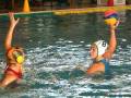 Dos jugadoras de waterpolo en acción, una con casco amarillo y otra con casco azul, luchando por un balón rojo y amarillo en una piscina.