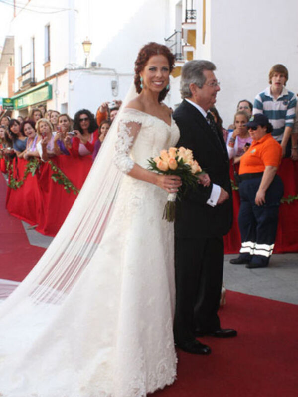 Novia en vestido blanco con velo largo y ramo de flores, caminando por alfombra roja en una celebración de boda.